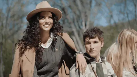 Friends in camel, plaid and knitwear layers smile and laugh while seated together outdoors, with trees behind them.