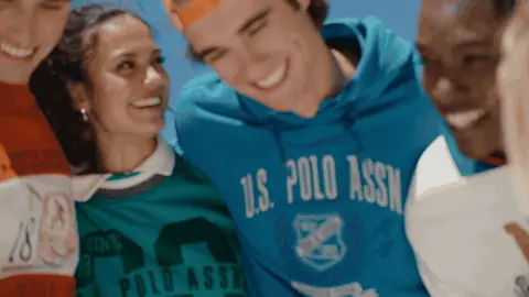 Friends in orange, green and blue sweatshirts lean shoulder to shoulder and laugh together on a sunny field.