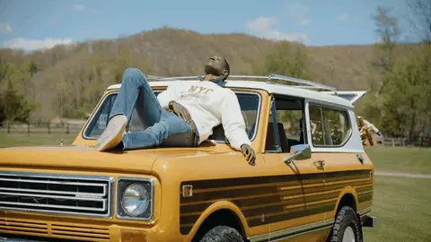 A man reclining on the hood of a yellow vintage SUV opens his arms and leans back in the sunshine.