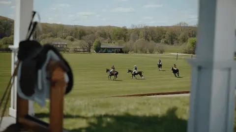 Polo players ride across a green field beyond white pavilion posts and a softly blurred saddle.