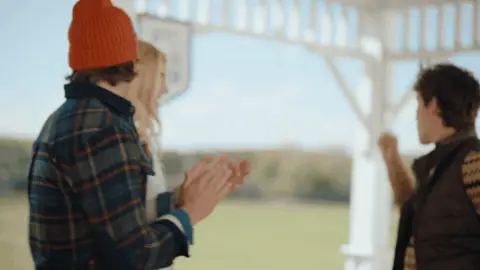 Friends in plaid, knitwear and quilted layers clap and smile while watching a polo match from a white pavilion.