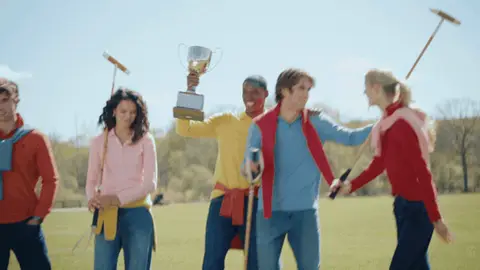 Friends in colorful polo shirts gather shoulder to shoulder with a raised silver trophy and wooden mallets on a green field.