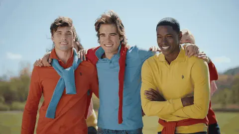 Two women jump onto their friends' backs as a group in colorful polo shirts laughs together on a sunny field.