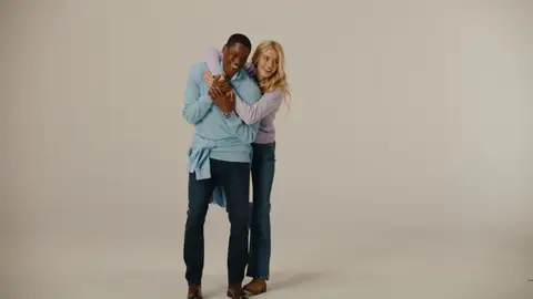 A woman in a lavender sweater hugs a smiling man in a pale blue sweater against a warm neutral studio backdrop.