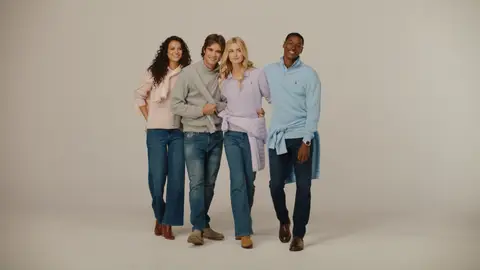Four adults stand close together in pink, gray, lavender and blue knitwear with jeans on a neutral studio sweep.