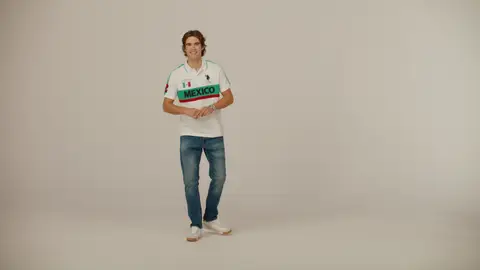 A smiling man holds his hands together, wearing a white Mexico-lettered polo with green and red bands, jeans and white sneakers.