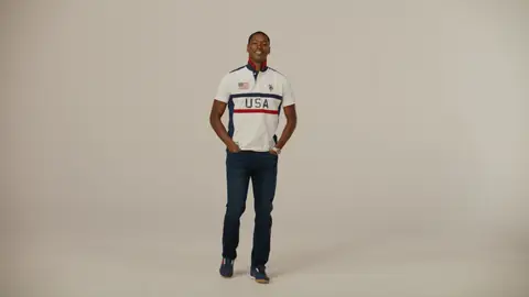 A smiling man wears a white USA-lettered polo with navy and red stripes, dark jeans and navy sneakers against a beige backdrop.