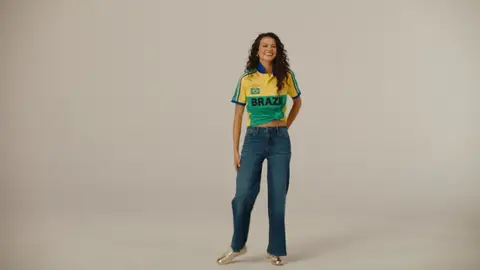 A smiling woman with curly hair wears a yellow and green Brazil-lettered polo, wide-leg jeans and metallic shoes against a beige backdrop.