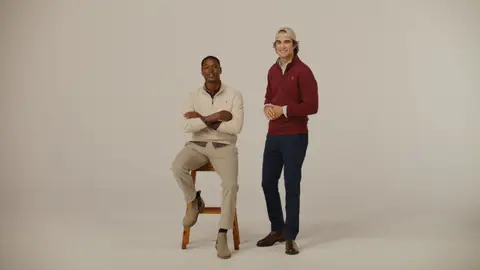 A man in cream knitwear sits on a wooden stool with folded arms beside a standing man in a burgundy sweater, navy trousers and a cap.