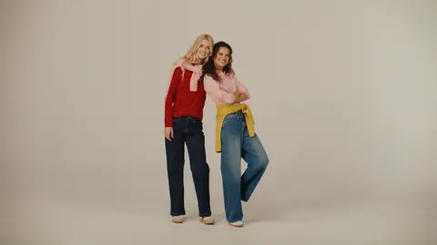 Two women lean together in red and pink sweaters and jeans, with a pink sweater around one neck and a yellow sweater tied at the other waist.