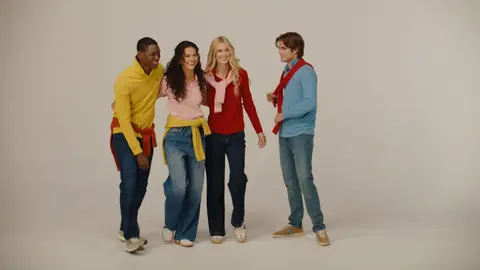 Four adults smile together in yellow, pink, red and blue knitwear with denim, against a warm neutral studio background.