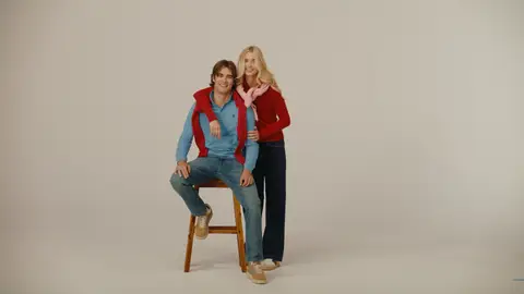 A man in a blue sweater sits on a wooden stool while a woman in a red sweater stands beside him with her arm around his shoulders.