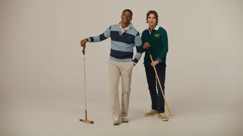 Two men hold polo mallets in a neutral studio, wearing a navy striped rugby shirt with cream trousers and a green top with navy trousers.