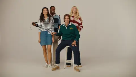 Four adults gather around a white stool in striped tops and a green pullover, with one man seated and a polo mallet resting behind the group.