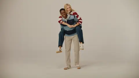 A smiling man in a navy striped rugby shirt and cream trousers carries a woman in a burgundy striped top and jeans on his back.