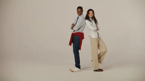 A man in a striped shirt, navy tie and jeans stands back-to-back with a woman in a pale shirt and beige trousers; a red sweater is tied at his waist.