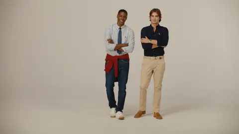 Two men pose with folded arms in striped and navy button-down shirts, paired with jeans and beige chinos on a neutral studio sweep.