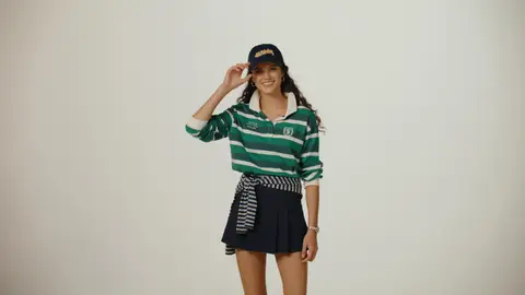 A smiling woman touches the brim of a navy cap, wearing a green-and-white striped rugby top, a navy pleated skirt and a striped sweater at her waist.