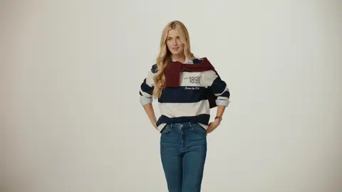 A blonde woman poses with her hands at her pockets, wearing a navy-and-white rugby shirt, jeans and a burgundy sweater around her shoulders.