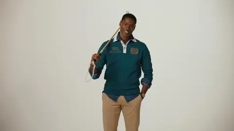 A smiling man in an embroidered teal polo and tan trousers holds a polo mallet over one shoulder against a neutral studio backdrop.