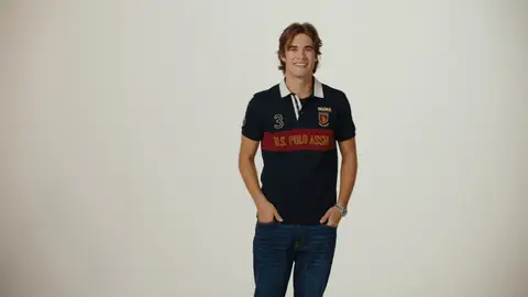 A man smiles with his hands in his pockets, wearing a navy polo with a red chest stripe and blue jeans on a pale studio background.