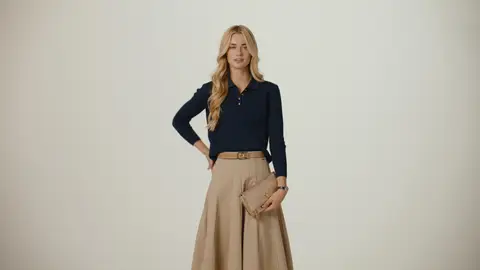 A blonde woman poses with one hand on her hip, wearing a navy knit top and tan pleated skirt while holding a tan clutch.
