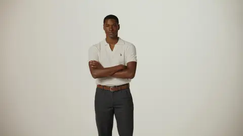 A smiling man folds his arms in a textured cream polo, dark gray trousers and a brown belt against a pale studio backdrop.