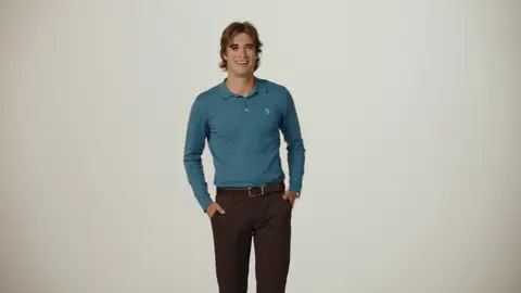 A smiling man stands with his hands in his pockets, wearing a blue long-sleeve polo and brown trousers against a neutral background.