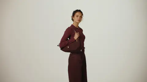 A woman turns in a burgundy long-sleeve polo and matching trousers, holding a burgundy chain-strap handbag over her shoulder.
