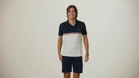 A smiling man wears a navy-and-white colorblock polo with a thin red stripe and navy shorts against a neutral studio backdrop.