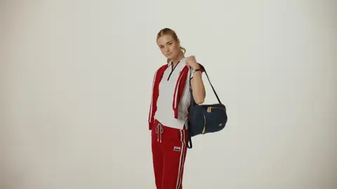 A blonde woman holds a navy bag at her shoulder, wearing a white polo, red track pants and a red layer draped over her shoulders.