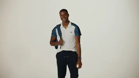 A smiling man wears a white polo with blue shoulder panels and navy trousers, carrying a navy backpack and holding a small white ball.