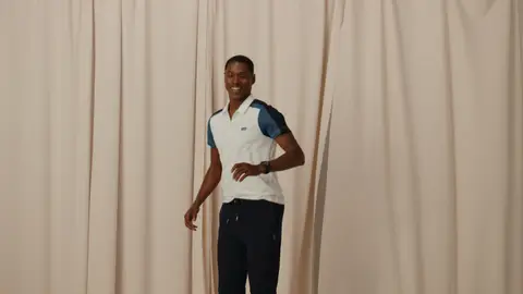 A smiling man turns toward the camera in a white polo with blue panels and navy trousers, framed by tall cream-colored curtains.