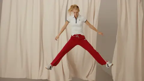 A woman jumps with her legs spread in red track pants and a white polo, suspended in front of flowing cream-colored curtains.