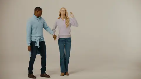 A man in a blue sweater raises their joined hands as a woman in a lavender sweater turns beside him in a neutral studio.