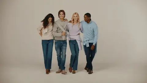Four adults in pastel sweaters and jeans take playful steps forward together against a neutral backdrop.