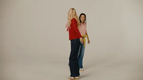 Two women in red and pink sweaters hold hands and turn together, with blue jeans and a yellow sweater tied at the waist.