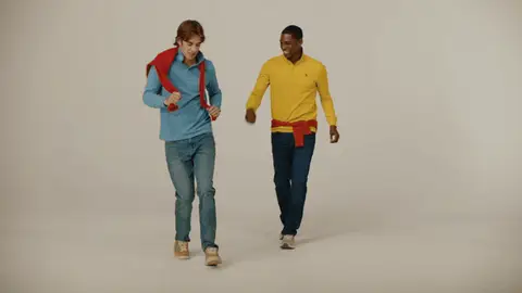 Two men in blue and yellow sweaters make playful dance steps on a studio sweep, with red knitwear tied around their shoulders and waist.