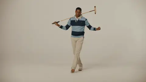 A man in a navy striped rugby shirt and cream trousers takes playful steps with a polo mallet resting across his shoulders.