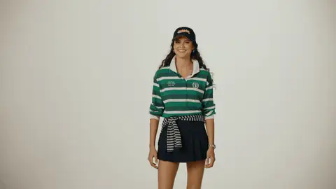 A woman in a green striped rugby top and navy pleated skirt touches the brim of her navy cap and smiles in the studio.