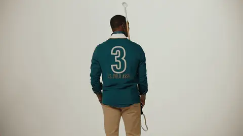 A man turns from the numbered back of his teal polo toward the camera while holding a polo mallet over one shoulder.