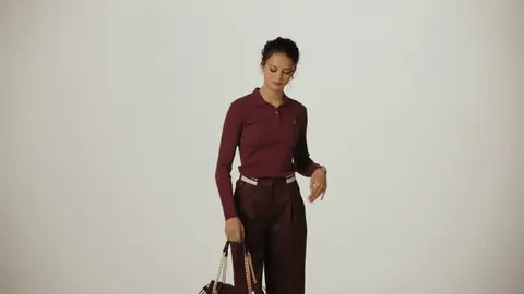 A woman in a burgundy polo and matching trousers lifts a chain-strap handbag onto her shoulder and turns toward the camera.