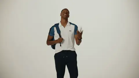 A man in a white and blue polo tosses and catches a small white ball while holding a navy backpack over one shoulder.