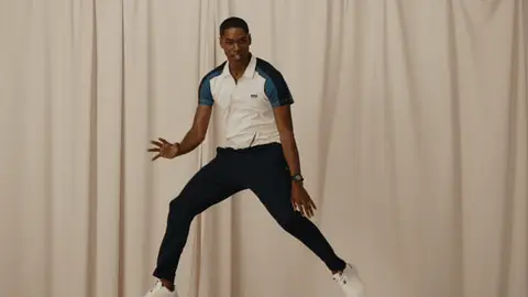 A man descends from a jump with one knee raised, smiling in a white and blue polo against cream-colored curtains.