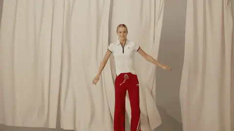 A woman in red track pants and a white polo springs into a wide-leg jump as cream curtains move behind her, then lands with a smile.