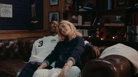 A woman in a navy sweater leans toward a smiling man in a white rugby shirt on a brown leather sofa.