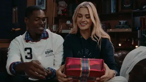 A man in a white rugby shirt and a woman in a navy sweater smile while holding a red gift with plaid ribbon on a leather sofa.