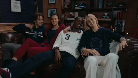 Four adults lean together on a brown leather sofa wearing navy, burgundy and cream casual clothing in a decorated room.