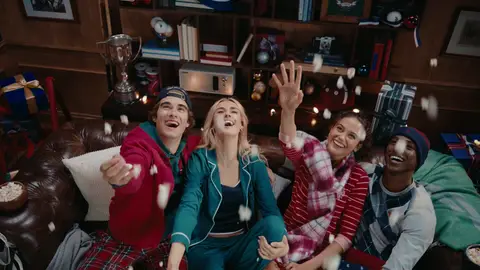 Four adults in colorful sleepwear look upward with raised hands as popcorn flies above a brown leather sofa.