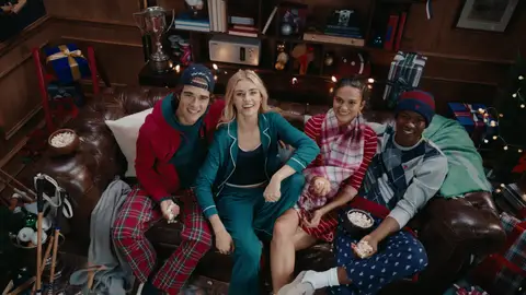 Four smiling adults in teal, red plaid and navy patterned sleepwear look up from a leather sofa beside wrapped gifts.